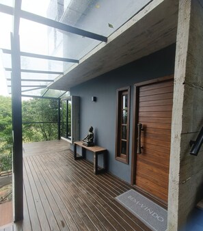 Una terraza de madera con un banco y una estatua de Buda, un muro de concreto y una puerta de madera con picaporte.