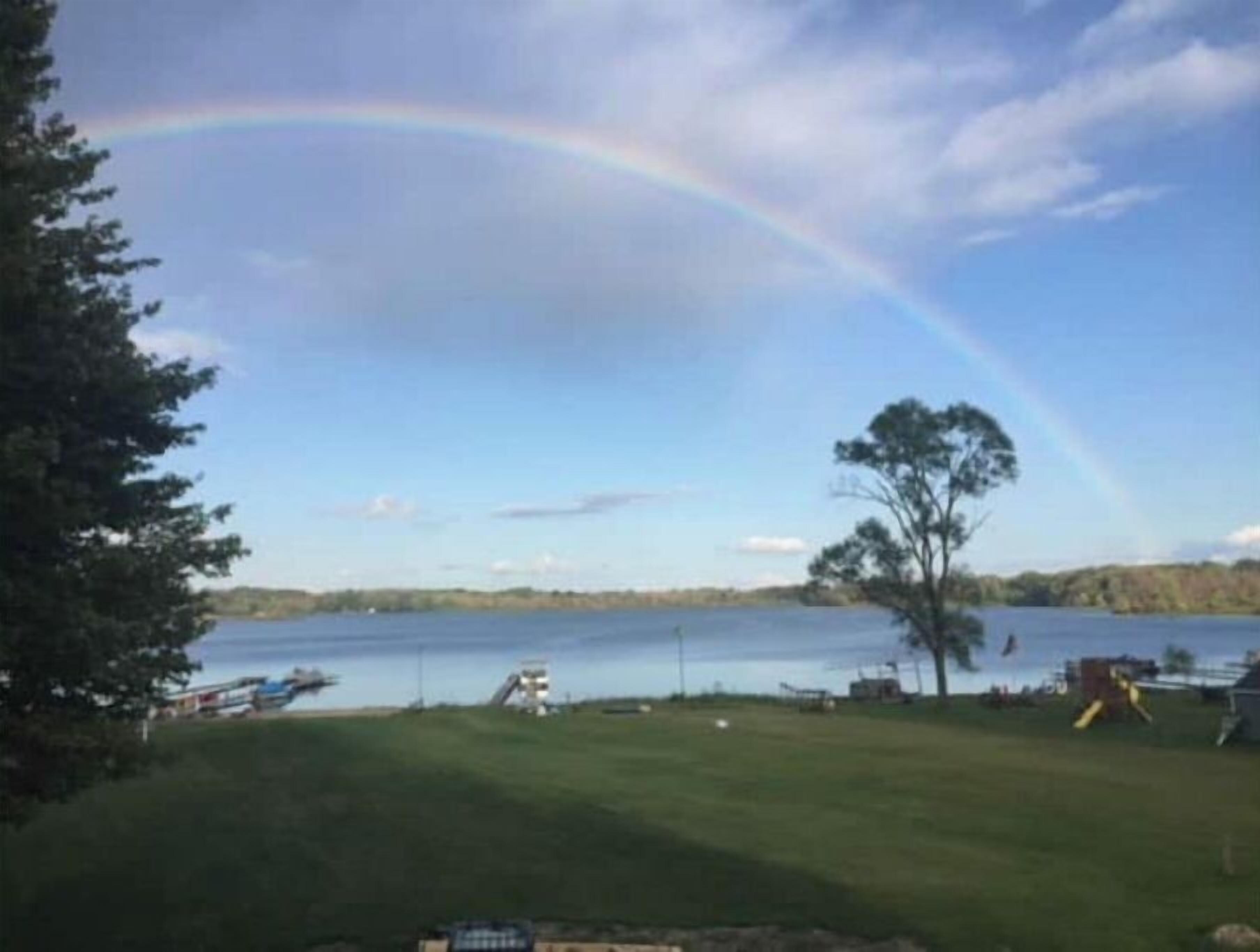A view of a lake with a rainbow, trees, and a grassy area with a playground.