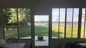 A room with a large window overlooking a grassy area and a body of water. There is a table with chairs, a small table with a flag, and a chair with a cushion. - Family Legacy Resort, Making great family memories (Allegan)