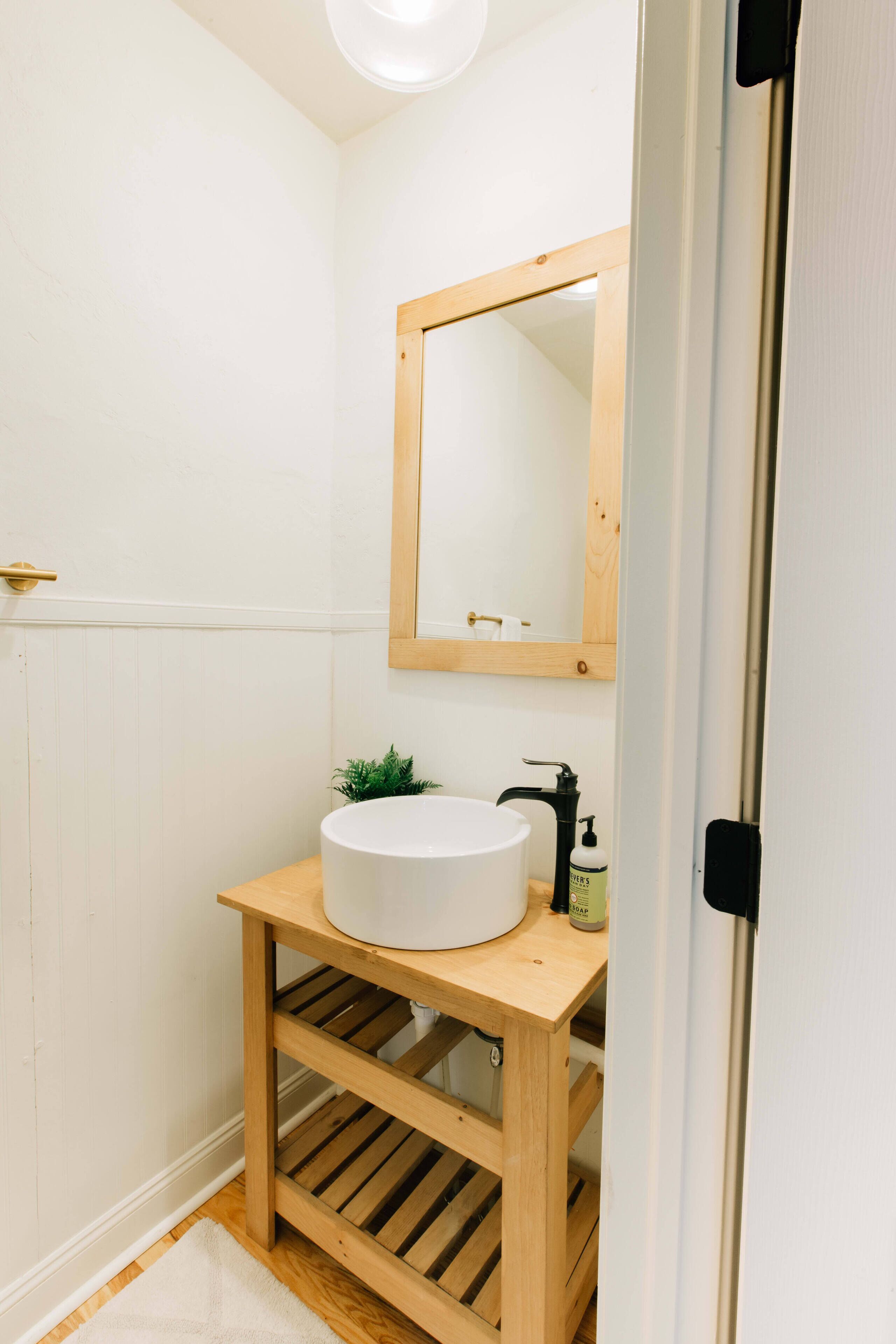 A bathroom with a wooden vanity, a white sink, a mirror, and a small plant.