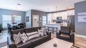 A modern living room with a sofa, armchairs, and a coffee table. There is a dining area with a table and chairs, and a kitchen with cabinets and appliances.