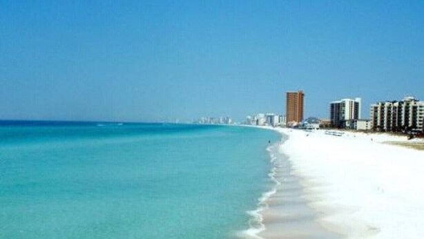 A beach with clear turquoise water and a sandy shore. There are several high-rise buildings along the coastline.