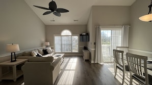A spacious living room with a large window, a ceiling fan, and a dining area with white chairs. - Endless Summer Updated Condo at Magnolia Place - Pool & Golf Course Views!  (Myrtle Beach)