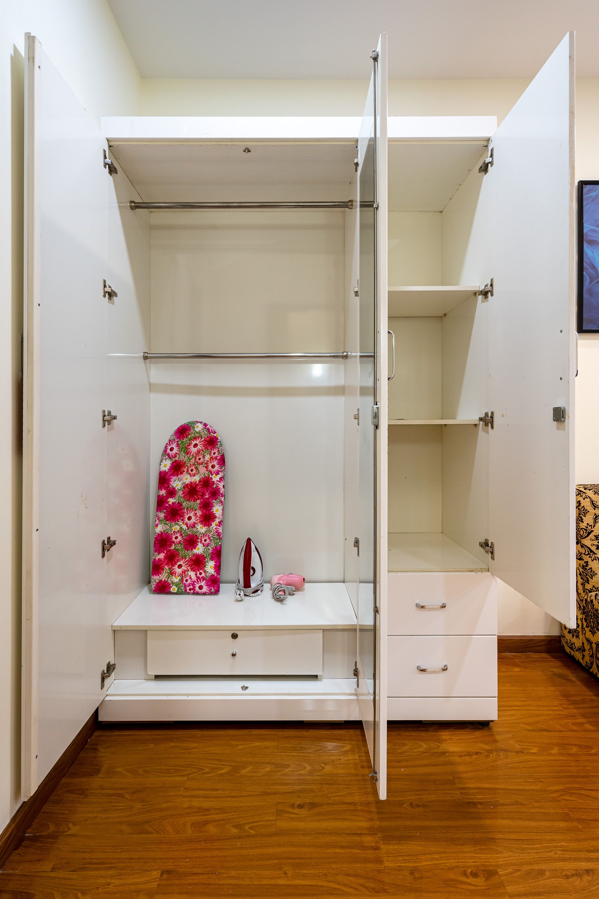 Um closet espaçoso com portas abertas, piso de madeira, prateleira com um objeto estampado floralmente e um pequeno ferro de passar.