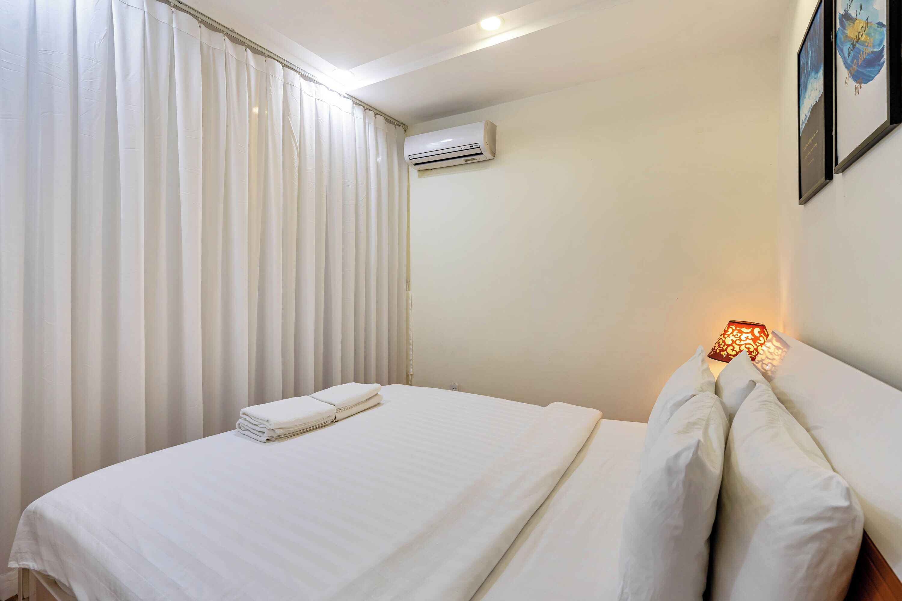 A neatly made bed with white linens, a wall-mounted air conditioner, and a bedside lamp.