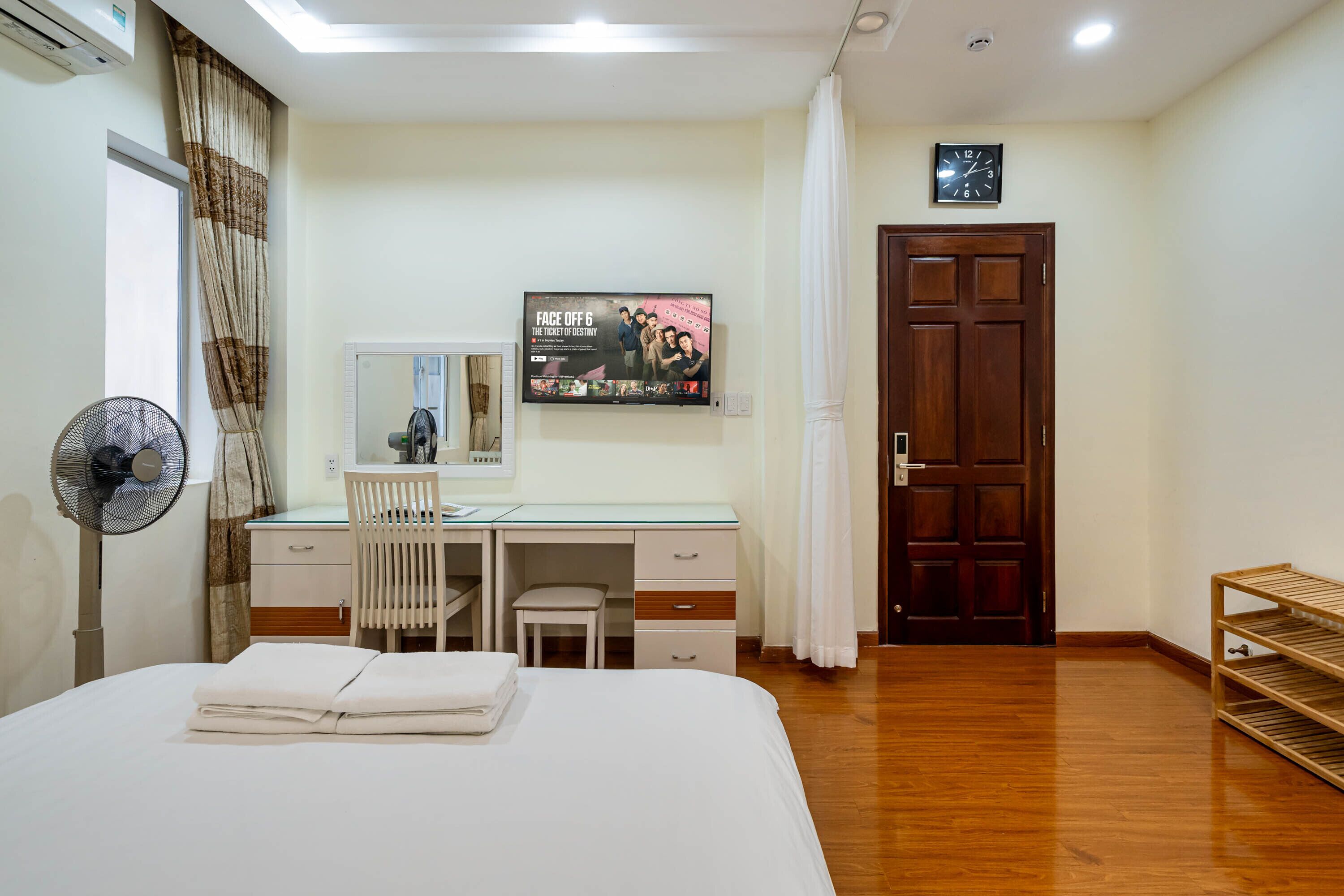 Une chambre d’hôtel avec un lit, un bureau, une chaise, un ventilateur, un miroir, une horloge et une porte.