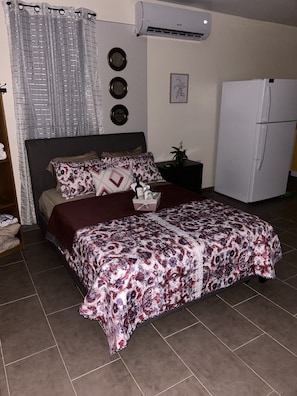 A bedroom with a bed, a nightstand, a white refrigerator, and an air conditioning unit.