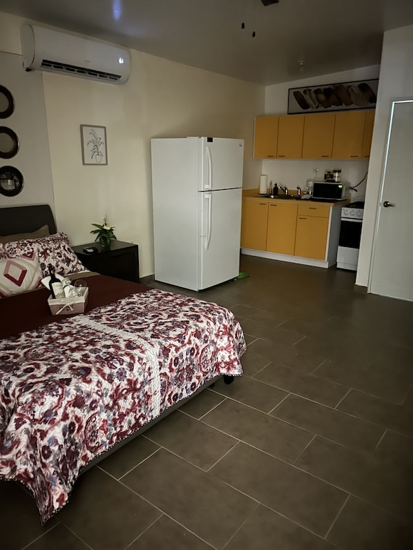 A bedroom with a bed, a nightstand, a white refrigerator, a small kitchen area with a microwave and stove, and a door leading to another room.