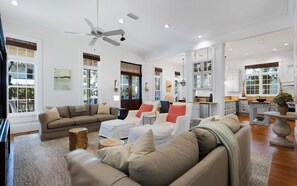 A spacious living room with a large sofa, a coffee table, and a dining area with cabinets and a kitchen counter. - WaterColor Home - Near Beach and Pools! (Santa Rosa Beach)