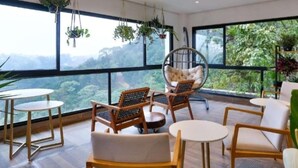Ein geräumiges Zimmer mit einem großen Fenster und Blick auf eine bewaldete Landschaft. Mehrere Holzstühle und ein runder Tisch, sowie ein Hängestuhl sind vorhanden.