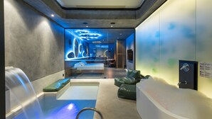 A modern spa room with a large bathtub, seating area, and a massage table.