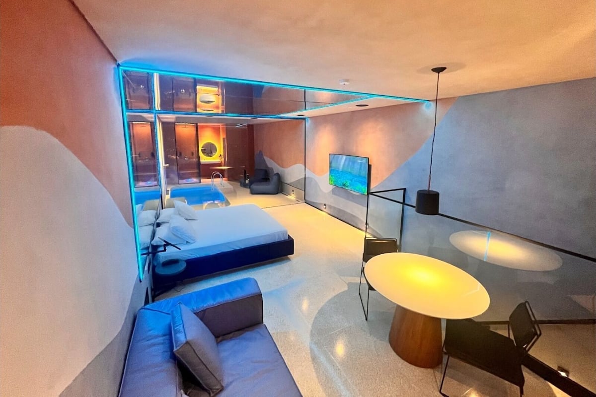 A modern hotel room with a glass partition, a bed, a sofa, a TV, and a small table.