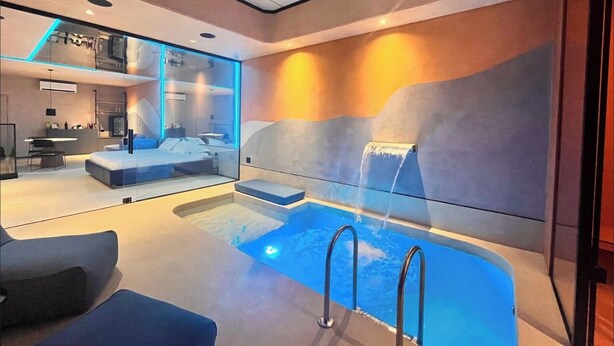 A modern hotel room with a large window, a built-in desk, a bed, and a small pool area.