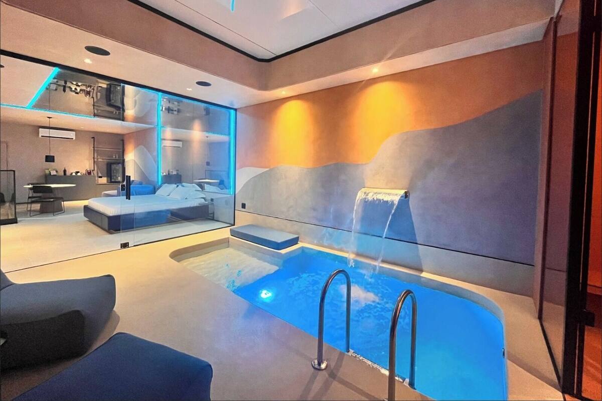 A modern hotel room with a large window, a built-in desk, a bed, and a small pool area.