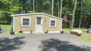 A small, single-story cabin with a beige exterior, a white door, and two windows. An American flag is displayed outside. - Nature Escape - studio in the forest (Middletown)