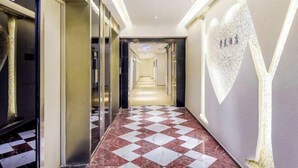 En hotellkorridor med ett rutmönstrat golv, hissar och en lång hall som leder fram till en ljusare del av byggnaden.