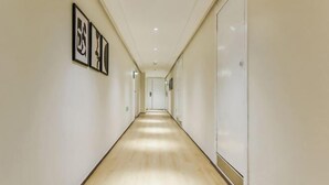 En lång, smal hall med ljusa väggar och ett trägolv. På vänster vägg hänger inramade tavlor och på höger vägg finns en vit dörr.