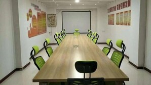 Un salón de conferencias con una larga mesa y sillas verdes, paredes blancas y una pantalla de proyector.