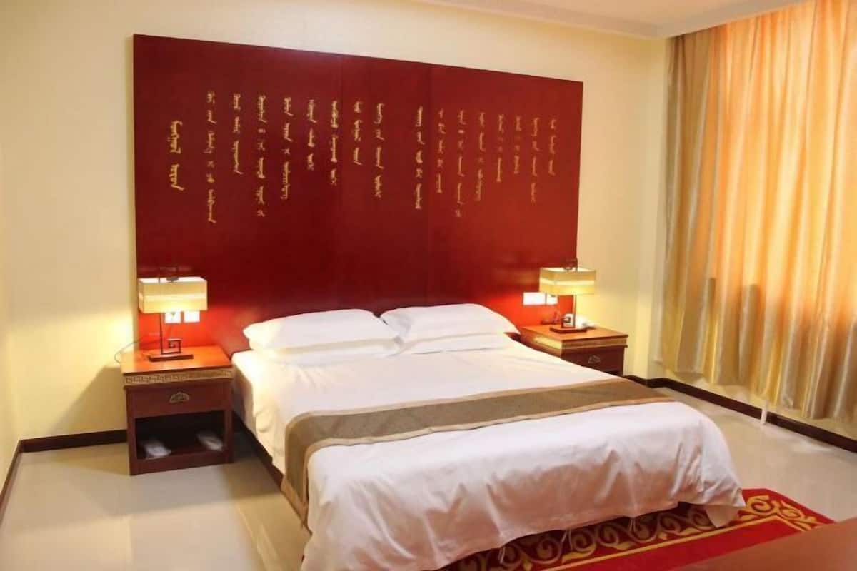 A hotel room with a large bed, two bedside tables, a red accent wall with gold script, and a window with curtains.