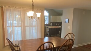 A dining area with a wooden table and chairs, a chandelier, and a view of the kitchen through a window with sheer curtains.