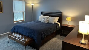 A bedroom with a dark blue bed, a bench, a nightstand with a lamp, and a window with blinds.