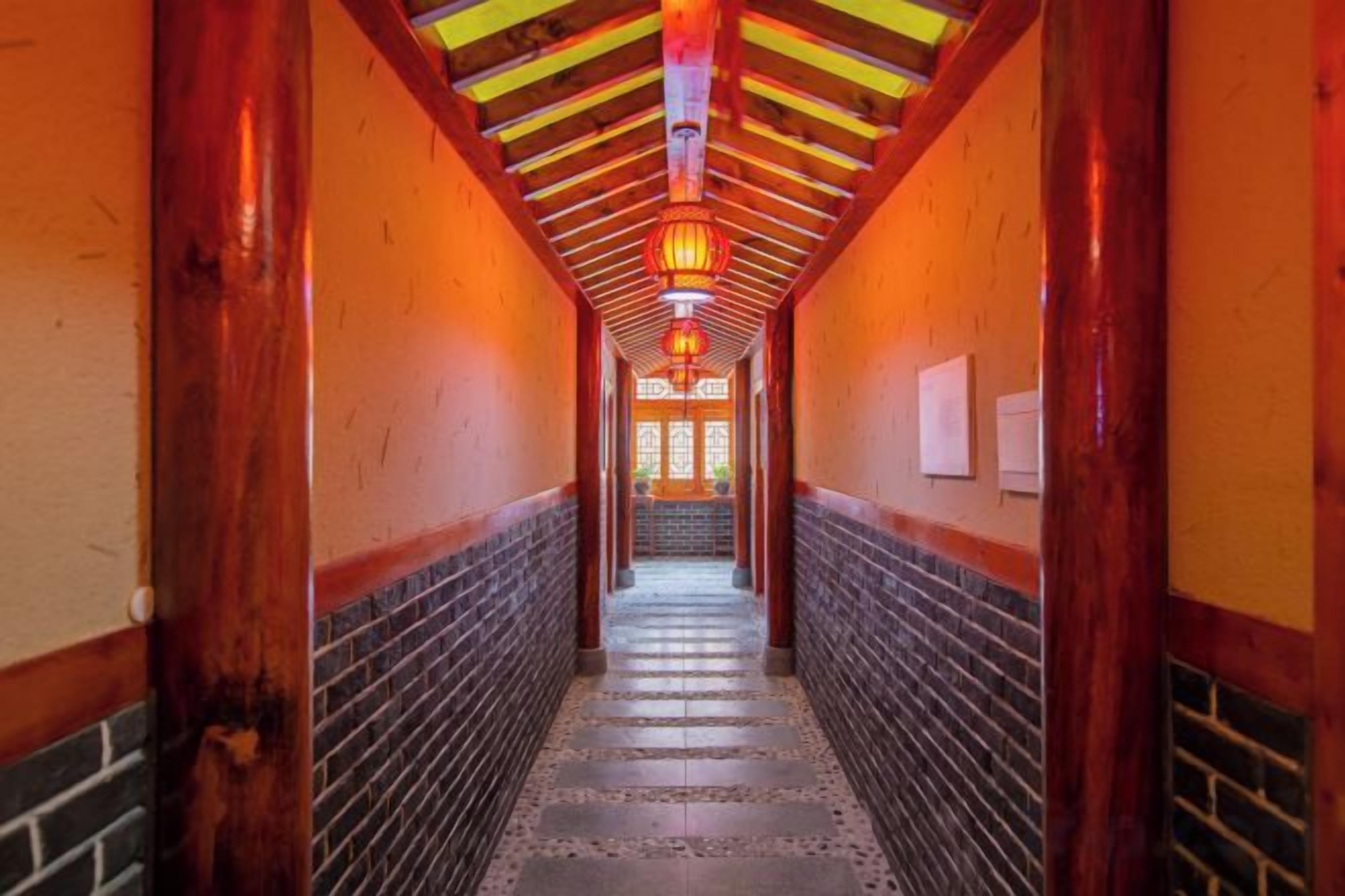 A corridor with wooden beams and a tiled floor, leading to a door with a red lantern.