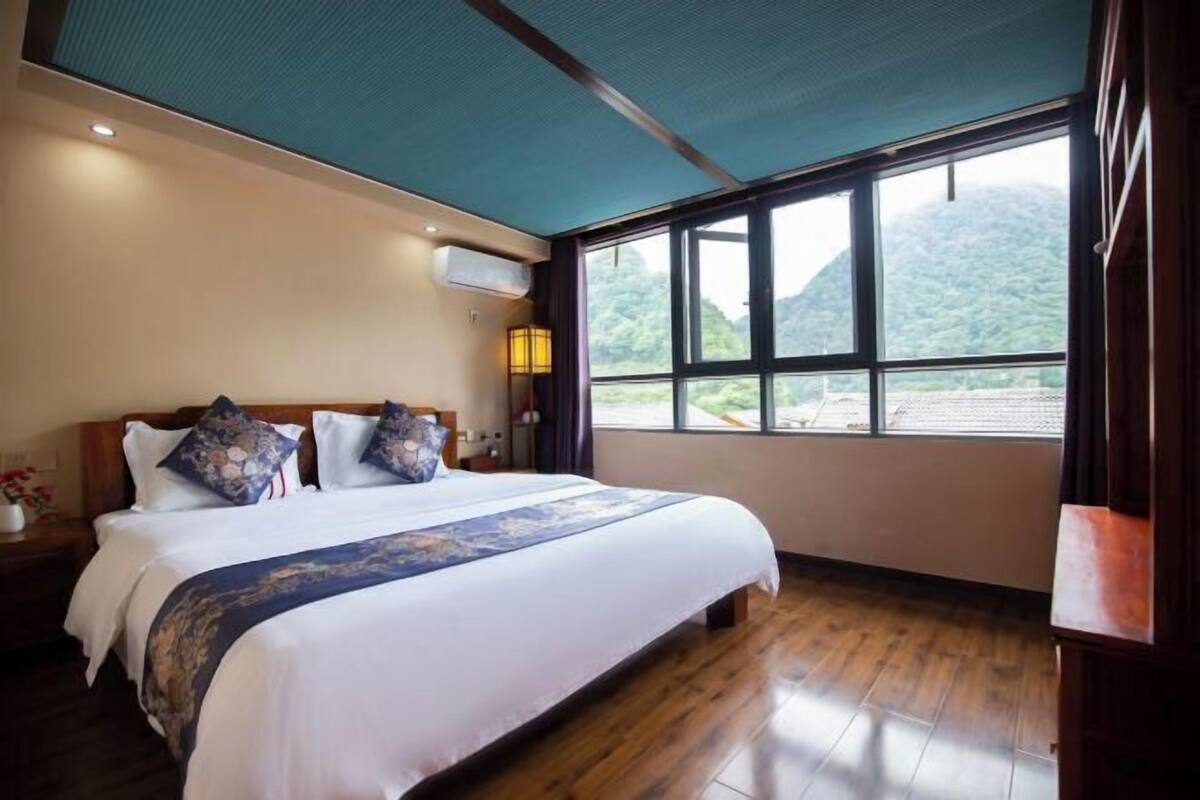 A bedroom with a large bed, a window with a mountain view, a ceiling fan, and a nightstand with a lamp.