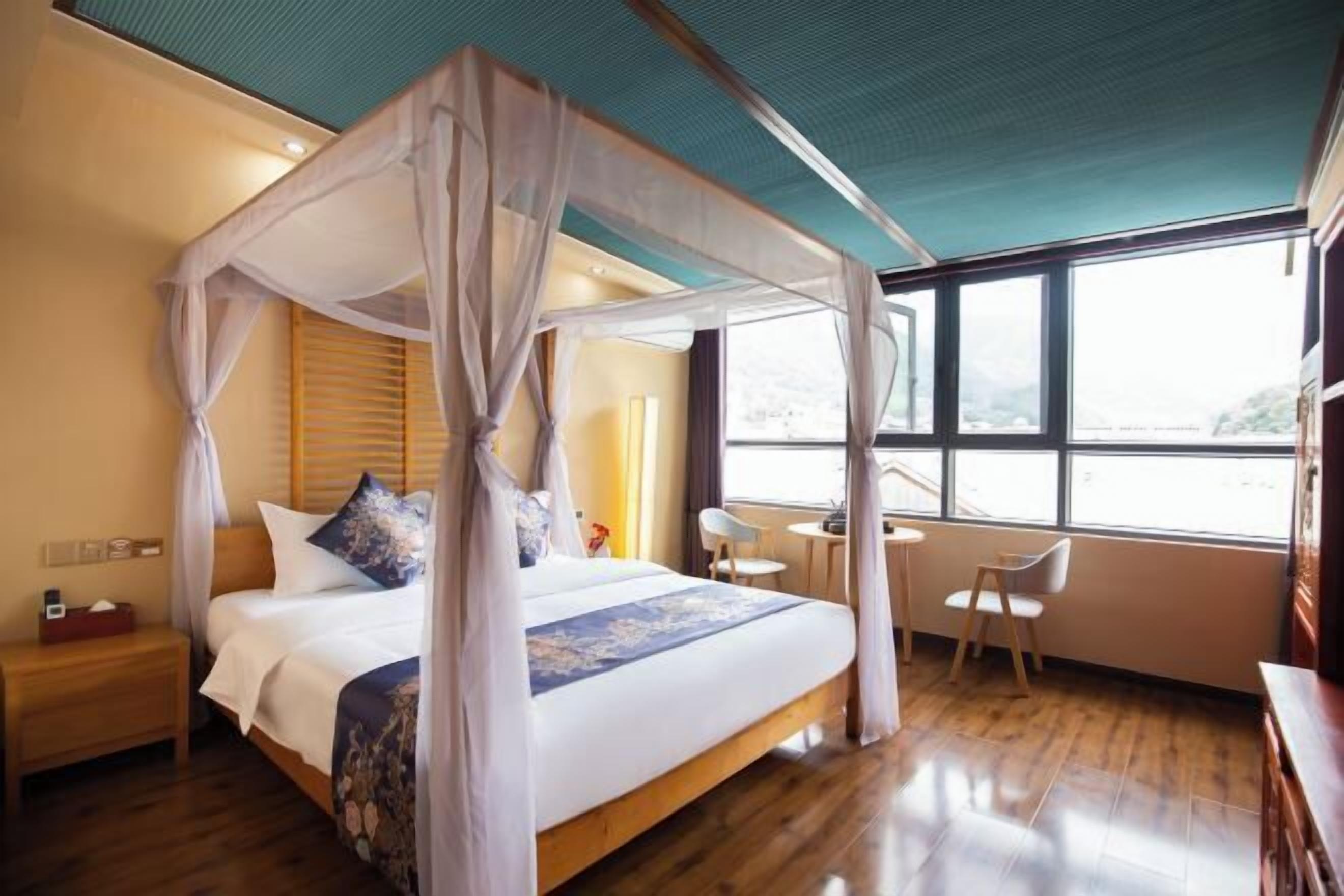 A four-poster bed with a canopy, wooden headboard, and a floral bedspread. There is a small bedside table with a lamp and a chair next to the bed. A window with a view of the outdoors is visible.