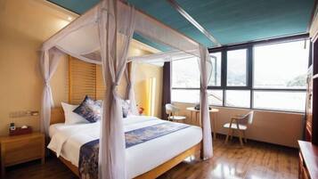 A four-poster bed with a canopy, wooden headboard, and a floral bedspread. There is a small bedside table with a lamp and a chair next to the bed. A window with a view of the outdoors is visible.