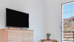 A minimalist room with a wooden cabinet, a television on top, a small table with a plant, and a large sliding glass door leading to an outdoor area. - Home2Book Comfy Apt Puerto Rico, Pool&Terrace (Mogán)
