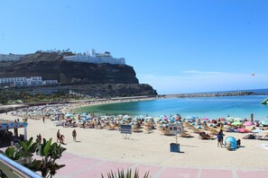 A beach with numerous colorful umbrellas and sun loungers, a clear blue sky, and a large building on a cliff in the background. - Home2Book Comfy Apt Puerto Rico, Pool&Terrace (Mogán)