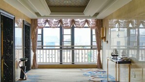Amplia habitación con ventanas grandes, alfombra estampada y una pequeña mesa con un modelo de barco.