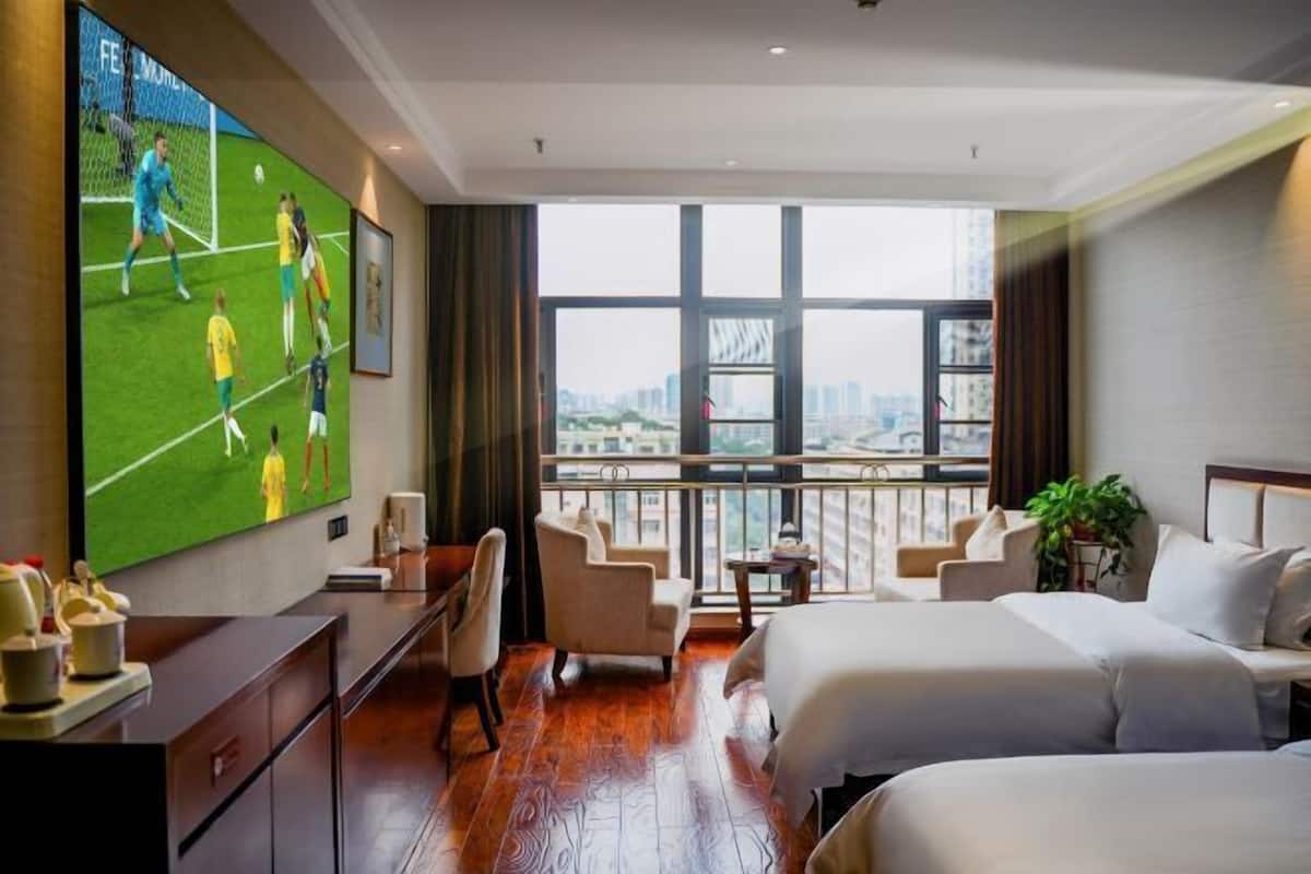Habitación de hotel con un televisor grande, un escritorio, dos camas, un balcón con vistas a la ciudad y un ventanal amplio.