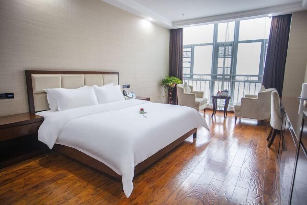Habitación de hotel con una cama grande, piso de madera, balcón con sillas y vista a la ciudad.