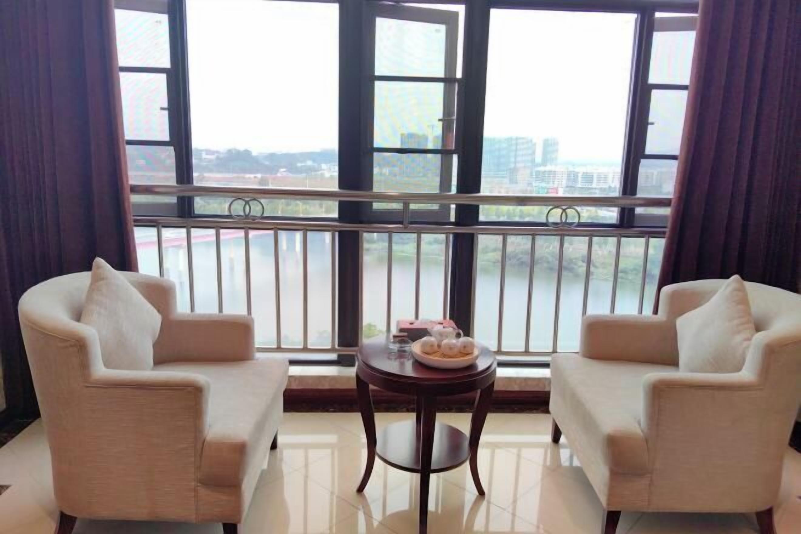 A room with two beige armchairs, a small round table with a bowl of fruit, and a view of a river and buildings through large windows.