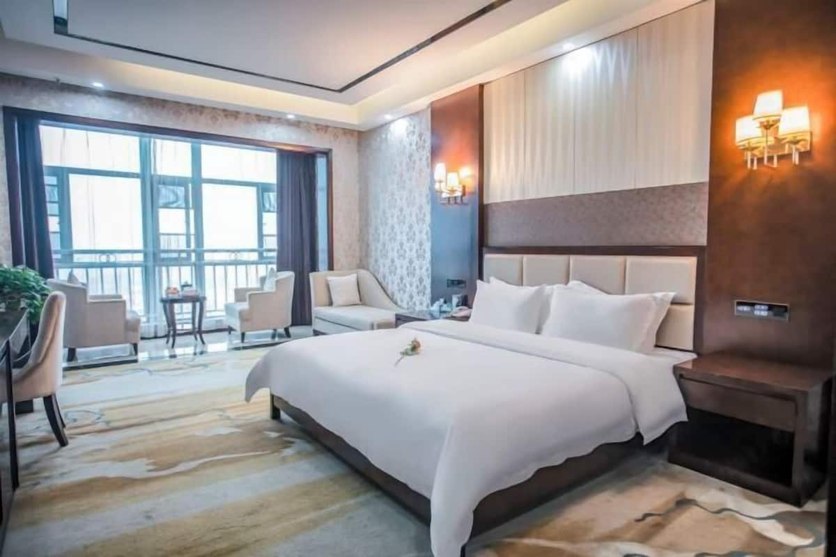 Habitación de hotel con una cama grande, un escritorio, una silla y una mesita. Se ve un ventanal con cortinas y vistas al exterior.