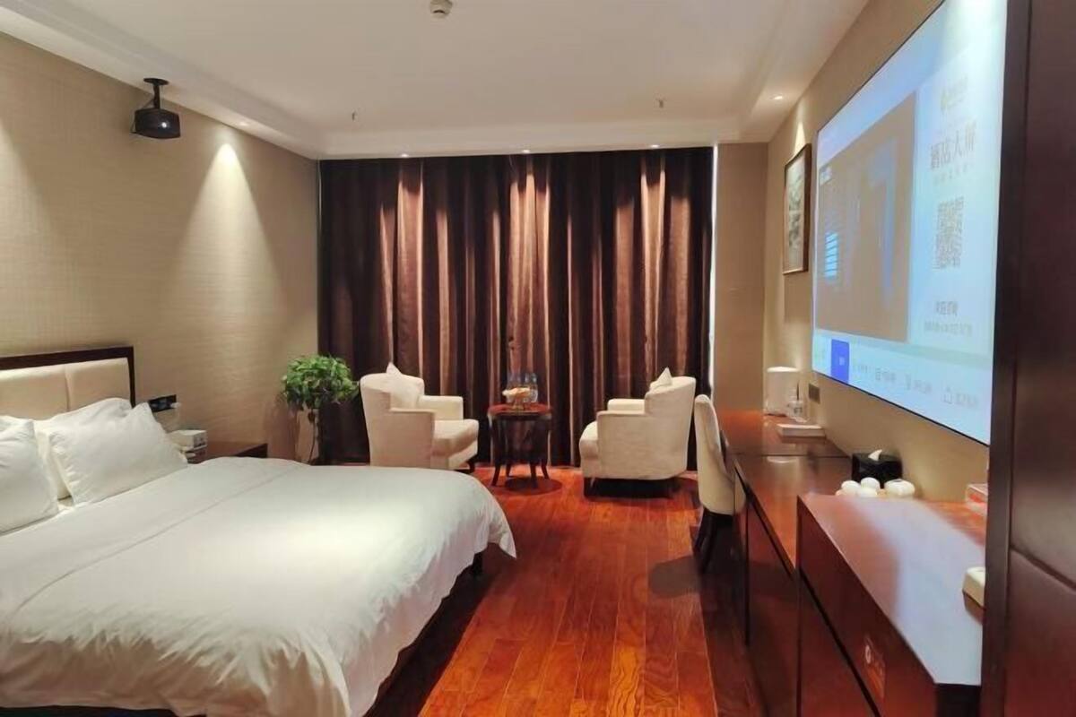 Habitación de hotel con una cama, dos sillones, una mesita, una planta y una pantalla de televisión.