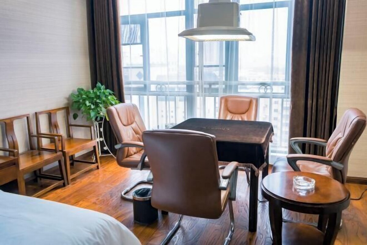 Una habitación de hotel con piso de madera, una cama, una mesa de comedor con sillas, una planta en maceta y un amplio ventanal con cortinas.