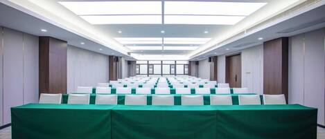 A conference room with rows of tables, a high ceiling with recessed lighting, and a view of the outdoors through the windows.