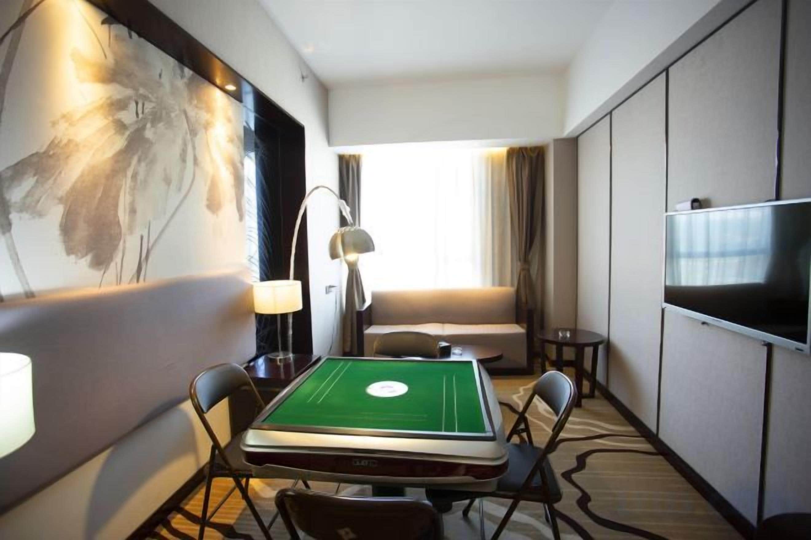 Habitación de hotel con una mesa de fieltro verde, un sofá, un televisor y un cuadro en la pared.