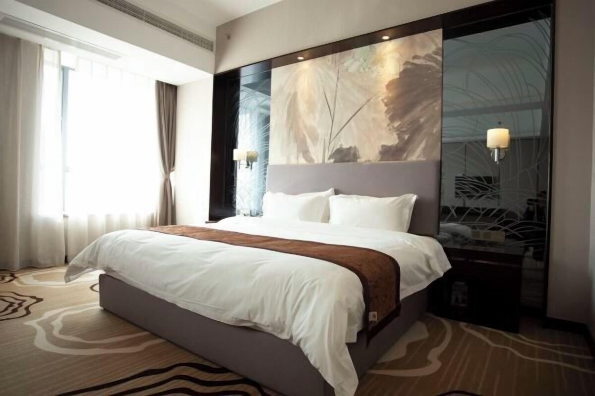 Une chambre d’hôtel avec un grand lit, une tête de lit ornée d’un panneau décoratif, une table de chevet avec une lampe et une grande fenêtre avec des rideaux.