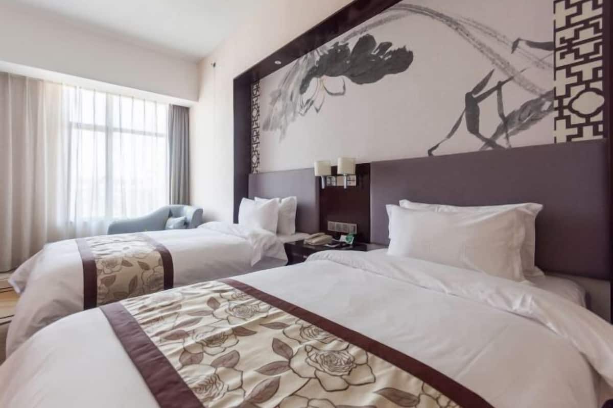 Camera d'albergo con due letti, un'ampia finestra con tende, una parete con un quadro e un comodino con una lampada.