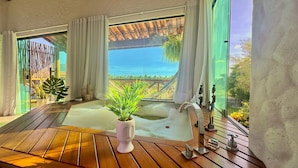 A room with a wooden floor, a large window with curtains, a white bathtub, and a potted plant.