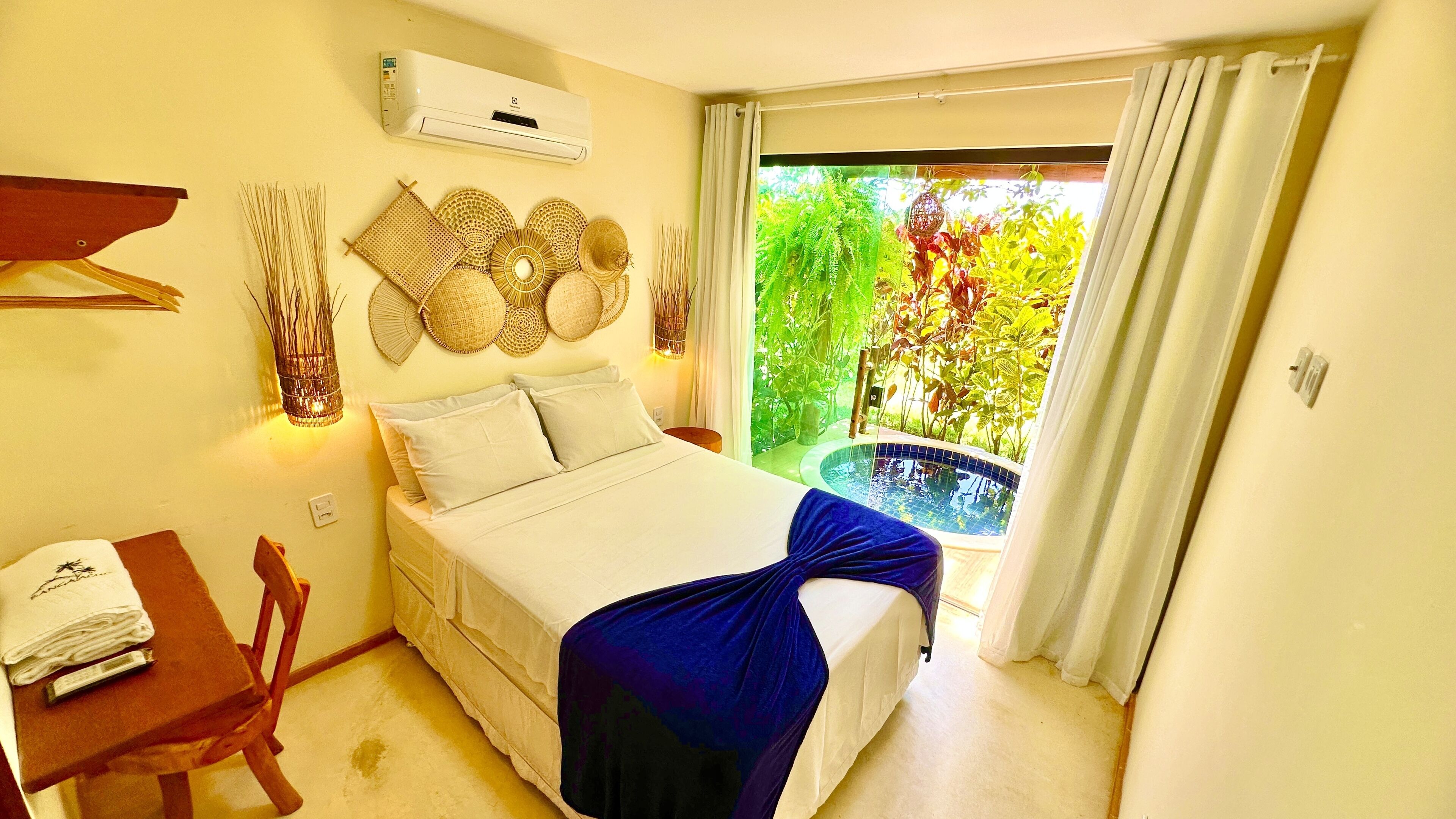 A bedroom with a bed, a small table with a towel, a chair, a wall-mounted air conditioner, and a view of greenery through a sliding door.