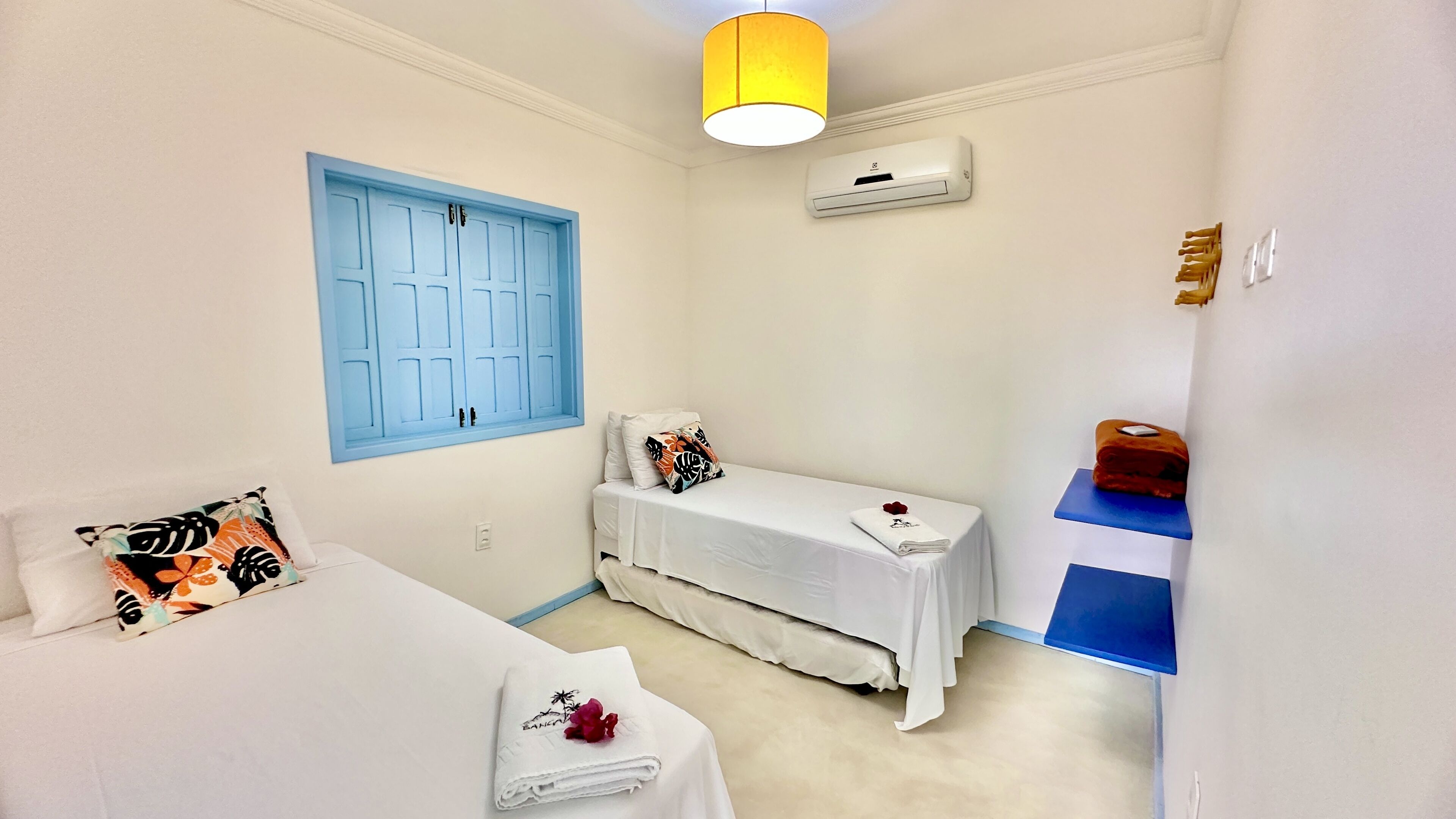 A small, clean room with a bed, a bedside table with a towel and a flower, a blue-framed window, and a wall-mounted air conditioner.