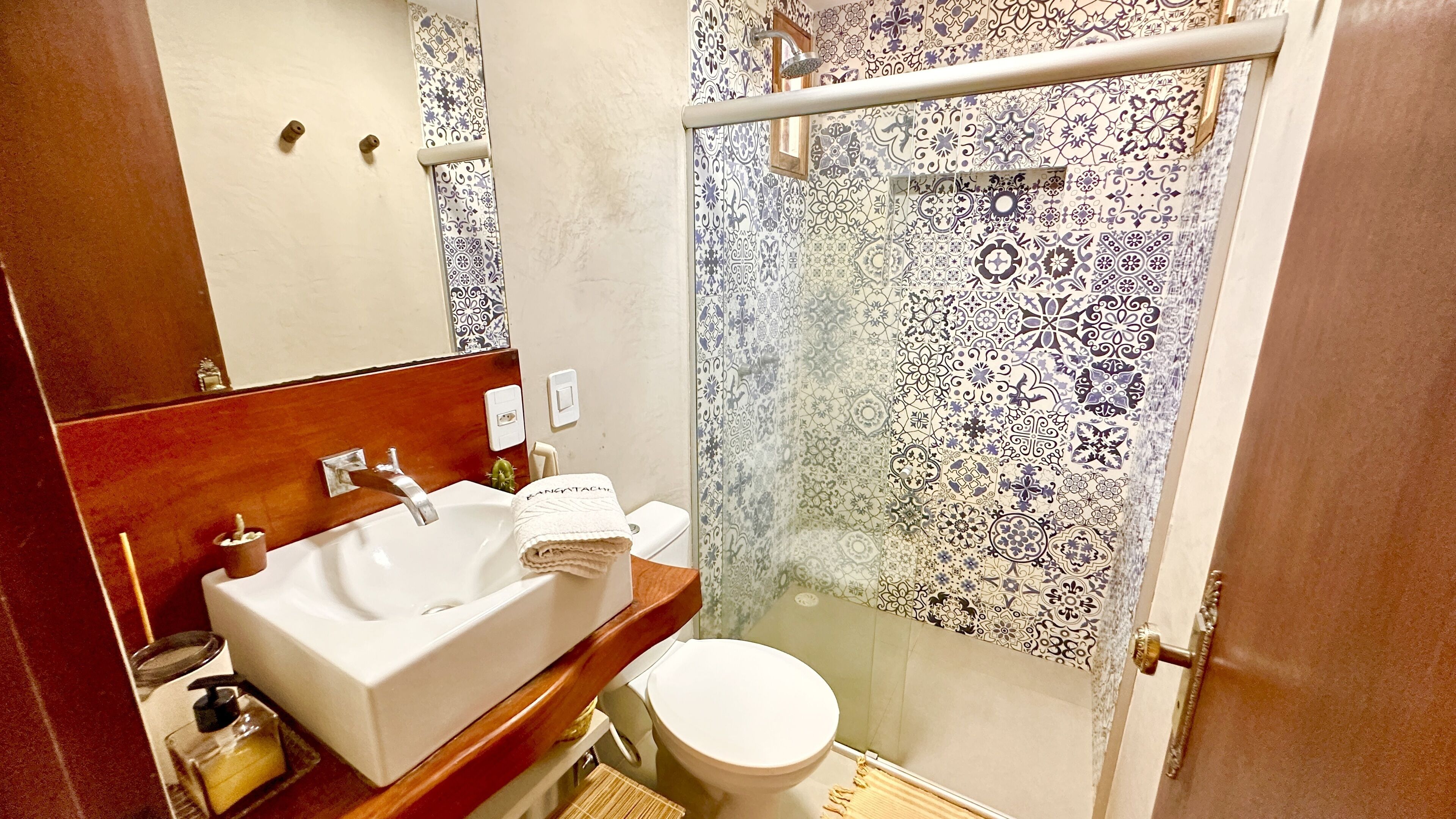 Un baño con lavamanos blanco, espejo, inodoro y ducha con azulejos estampados.