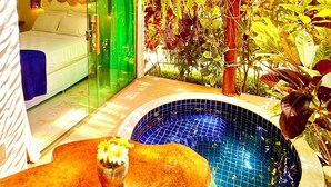 A room with a glass door leading to a bathroom, a wooden ceiling, and a small pool with a wooden table and flowers in the foreground.