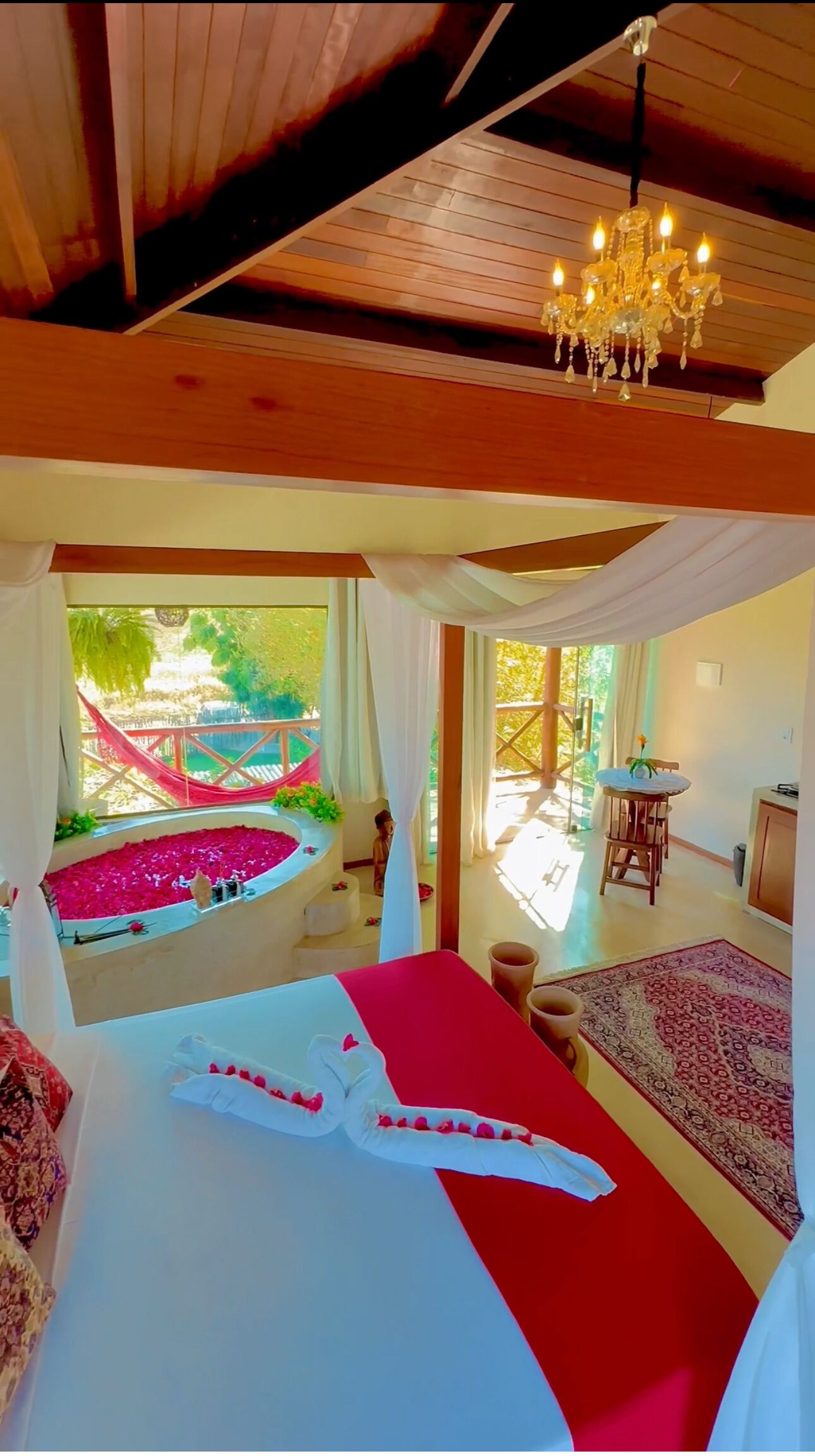 Een slaapkamer met een groot bed, een kroonluchter, uitzicht op een tuin, en een zithoek met een tafel en stoelen.