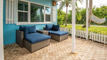 A covered outdoor seating area with wicker furniture and blue cushions, a white picket fence, and a hammock in the background.