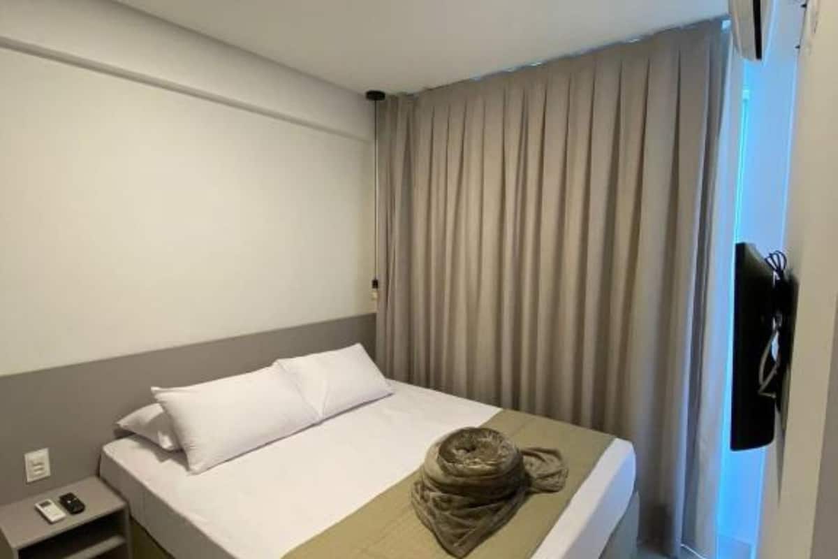 A hotel room with a bed, a bedside table, a wall-mounted air conditioner, and a window with curtains.